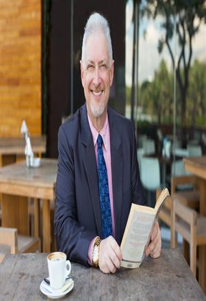 Closeup portrait of smiling senior handsome man looking at camera, reading book, drinking coffee and sitting at table in outdoor cafe. Front view.